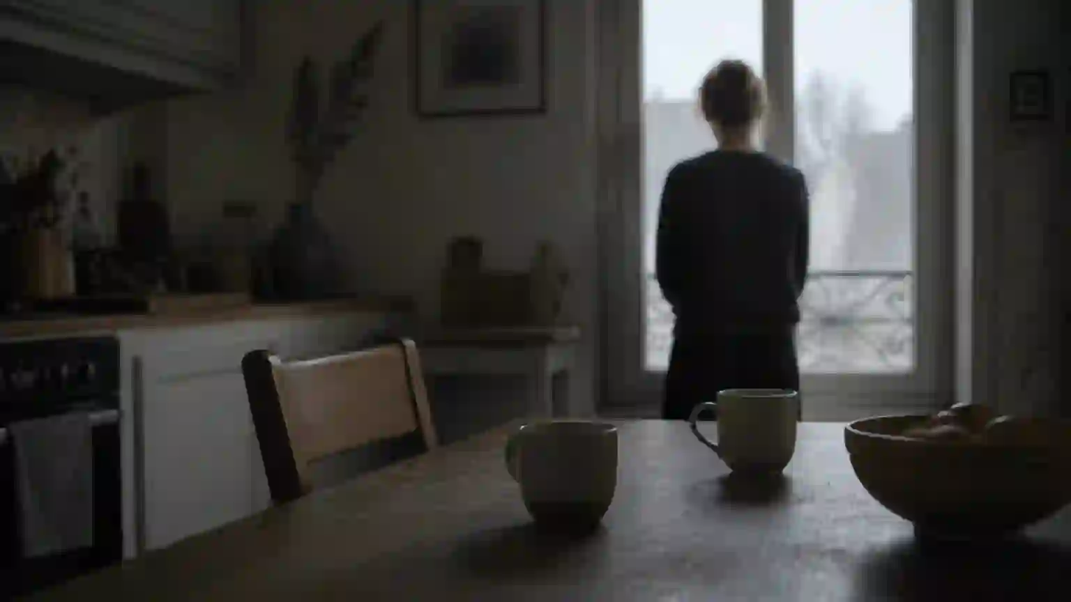 Tôt le matin dans une cuisine ordinaire : deux tasses posées sur la table de bois, une chaise écartée, et au fond, de dos et floue, une personne immobile devant la fenêtre.